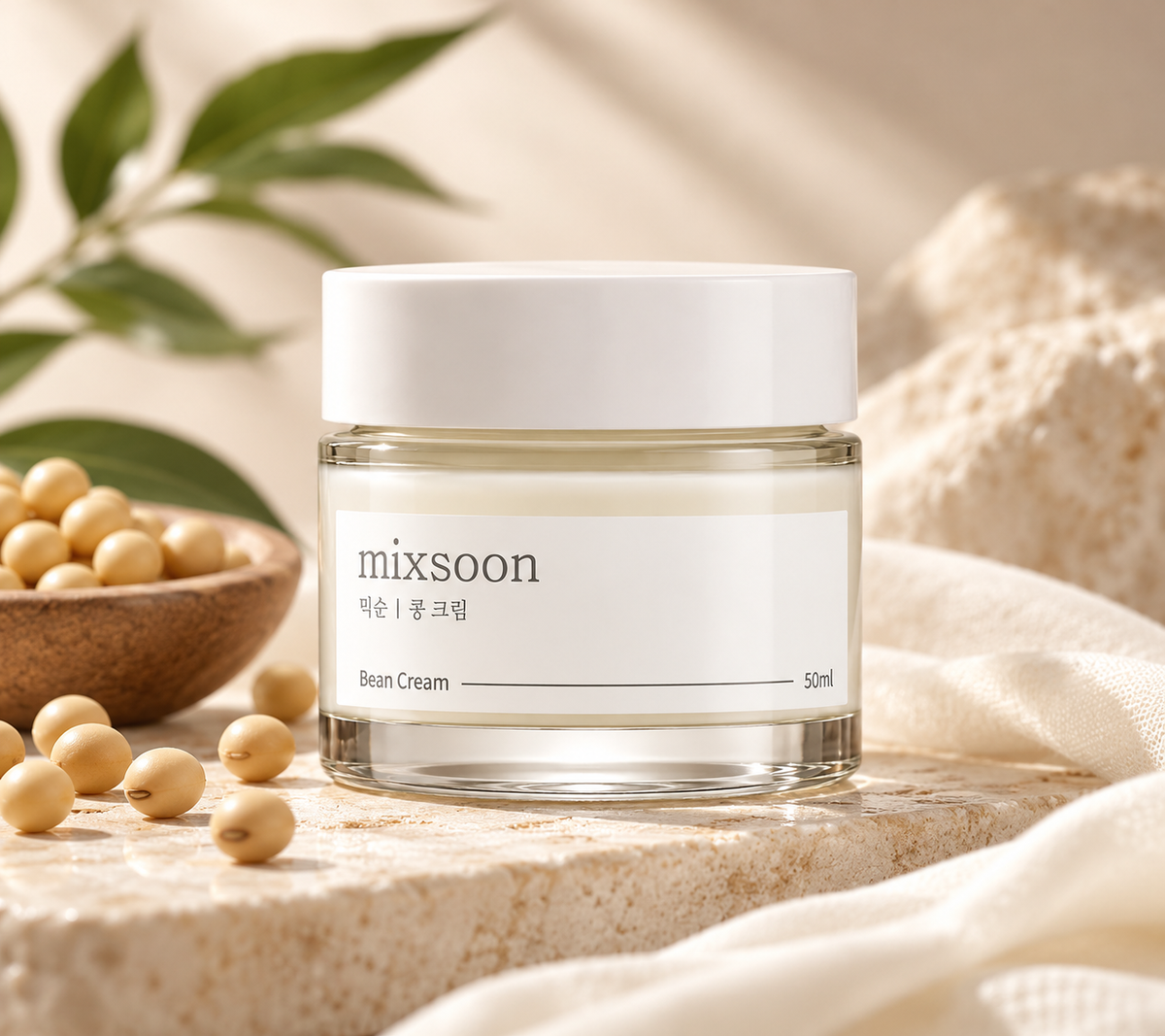 Mixsoon Bean Cream 50ml Korean skincare moisturizer in an elegant glass jar, surrounded by soybeans, fresh green leaves, soft ivory fabric and natural stone. A hydrating facial cream presented in a luxurious minimalist setting with warm natural lighting, highlighting gentle Korean skincare and daily skin hydration.