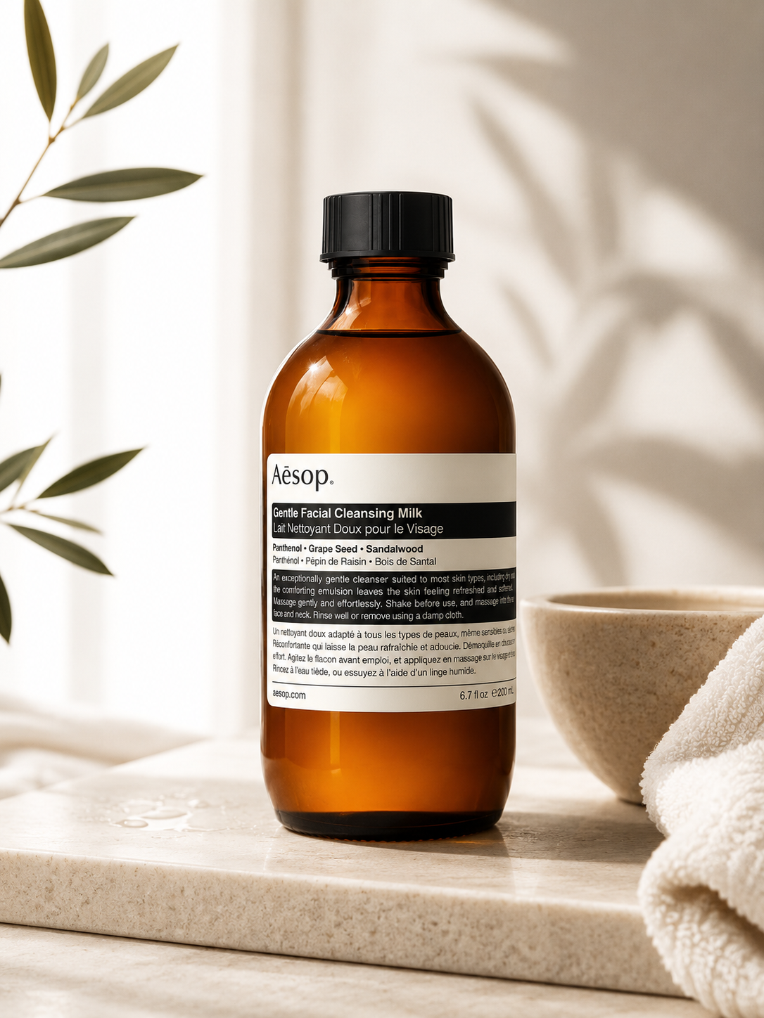 Aesop Gentle Facial Cleansing Milk displayed on a natural stone countertop in a serene, minimalist skincare setting with soft natural light, botanical greenery, a ceramic bowl, and a folded white towel. The warm, spa-inspired atmosphere highlights the amber glass bottle and reflects the cleanser's soothing, nourishing formulation for gentle daily facial cleansing.